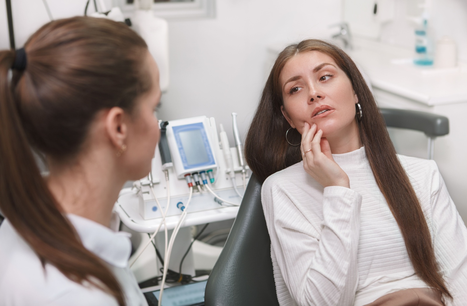 A person sitting in a dental chair holds her jaw while talking to the dentist about her TMJ